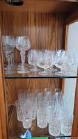 Top and middle shelves of a wooden display cabinet containing multiple etched crystal glasses of different types.
