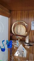 Photo showing the brass dinner gong with wooden striker, two glass bird figurines, decorative ship model, and glass pyramid on display shelf.