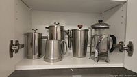 Shelf with multiple coffee and tea accessories including stainless steel French press coffee pots, glass French press, frother, and pewter tankard.