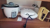 Photo of the lot on a wooden shelf showing a white Black & Decker rice cooker with glass lid, a white George Foreman grill, and a beige GE hand blender with beaters attached.