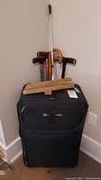 Photo showing one closed large black Outbound suitcase with several walking canes and a wooden boot remover leaning against the wall