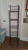 Front view of a tall, narrow metal shelving unit with five wire shelves and decorative metal on top shelf back.