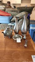 Pewter teapot, two tall pewter vases, smaller pewter pitcher/bud vase, small round pewter dish, and collector spoon in packaging all arranged on wooden table.