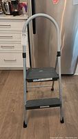 Front view of the Cosco folding step stool showing two black plastic steps and a curved metal handle.