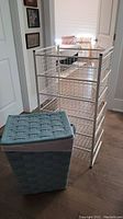 Photo showing the white metal storage stand with five wire drawers and the blue fabric laundry hamper with lid placed in front of it.
