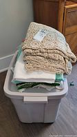 Stack of various table linens including crocheted tablecloths on top and mixed fabric linens below in plastic storage bin.
