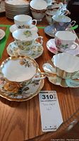 Photo showing six teacup and saucer sets arranged on a wooden surface, showcasing floral patterns and gold trims