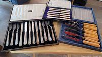 Photo showing three flatware sets including silver plate knives and forks, Maitland's white bakelite handled seafood knives in a box, and 6 plastic handled knives and forks.