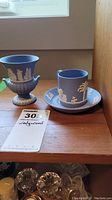Photo of Wedgwood 3.5 inch urn and demi tasse cup with saucer on wooden shelf showing classical white relief decoration on blue jasperware pieces.