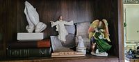 Shelf with multiple religious and decorative figurines including praying hands, angels and a glass bell. A Bible and other books placed underneath.