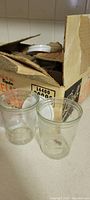 Two small clear glass jelly jars in front of vintage Ball Mason jars box containing lids visible inside.