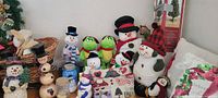 Collection of snowman figurines in various styles and sizes, with two green frog plush toys, assorted mugs, and a decorative pillow in the background.