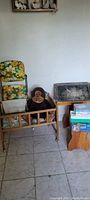 Wooden doll bed with a stuffed monkey inside, adjacent to old high chair with yellow floral seat upholstery, and wooden play school desk showing chalkboard surface.