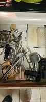 Top view of a kitchen drawer showing mixed metal and plastic kitchen utensils including graters, peelers, tongs, scissors, and a meat tenderizer