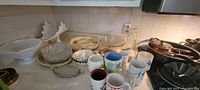 Overview of assorted kitchen dishes, bowls, mugs, and pans on a counter including white ceramic dishes, glass bowls, floral mugs, and stacked black pans.