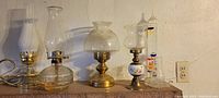 Collection of five electric, oil, and candle lamps with various glass and brass bases, plus a Galileo thermometer on the far right.