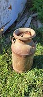 Rusty vintage metal milk container with handle and lid sitting on grass outdoors.