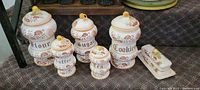 Five ceramic kitchen canisters with floral design labeled Flour, Sugar, Coffee, Cookies, and Tea with round yellow knobs on lids.