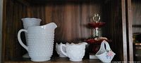 Milk glass set including hobnail pitcher, creamer, bowl, and a two-tier red glass serving dish, all displayed on a wooden shelf.