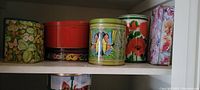 Photo showing seven assorted decorative tins on a shelf, including floral patterns, red, green tins and butterfly artwork.