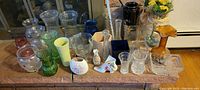 Wide view of multiple flower vases arranged on a brick ledge showing different shapes and colors including clear, green, yellow, blue, amber, and floral decorated vases.