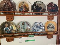 Full view of eight collector's plates arranged on two levels of a wooden display shelf, showcasing varied artwork and plate shapes