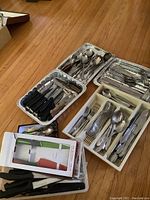 Overall view of cutlery and knives arranged in trays and boxes on wooden floor, showing variety of pieces and containers.