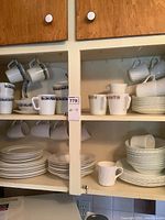 Open white kitchen cabinet holding assorted dishes and mugs with blue floral and gold patterns hanging and stacked on shelves.