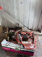 Photo showing box with tangled electrical supplies including orange extension cords, power bars, and a green trouble light inside a cardboard box labeled 'Sweet Broccoli Tender Stems'.
