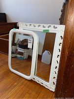 Both white wood framed mirrors shown leaning against wall on hardwood floor with some household items reflected in the glass.