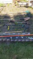 Photo showing various garden hand tools including shovels, rakes, a garden fork, and levels arranged on the ground.