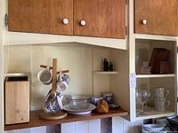 Shelf with blue and white mugs hanging, ceramic bowl and muff in front, wine glasses in cabinet, wooden shoe, and small dark figurines visible