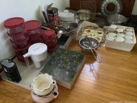 Wide view of the lot showing multiple stacked Pyrex bowls with red lids, clear glasses with floral designs, divided ceramic serving tray, apple peeler/corer, and assorted mugs.