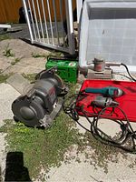 Overall view of all tools laid outside on concrete and grass; bench grinder, charger, drills, sander visible.