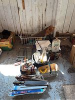 Photo showing various saws including wood handled hand saws arranged in a basket, with other small tools, oils, and boxes around on a rough floor.