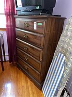 Image showing front view of five drawer wood highboy dresser with brass handles, an older model JVC TV on top, and an ironing board next to it.