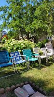 Photo shows six lawn chairs arranged on grass with greenery background including one folding wooden chair with tan cushion, two white resin armchairs, and three metal folding chairs with blue, light blue, and teal webbed seats.