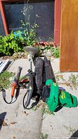 Full view of all three garden tools laid on concrete: green electric blower/vacuum, grey and orange weedwacker, silver and black hedge trimmer