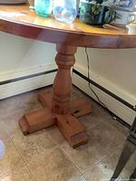 Close up of the four-legged wooden pedestal base supporting the round pine table top on tile flooring, showing the mostly sturdy structure and wood finish.