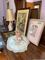 Photo shows four framed prints including one with floral and one figurative artwork, porcelain doll in white dress, and ceramic base lamp with floral design and beige shade, all displayed on wooden chest.