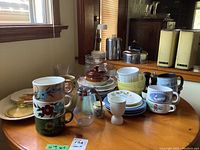 Wide view showing stacked soup mugs with floral designs, mixed plates on the table, salt and pepper shakers, and glass salad set.