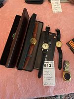 Wide shot of all five men's watches, some in cases, on a pink surface with lot information card