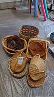 Eight woven baskets displayed on floor, one containing pinecones, showing variety in shapes and weaving styles.
