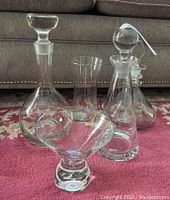 Photo showing three clear crystal decanters with stoppers and two glass vases placed on a red carpet with a grey couch background.