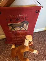 Photo of red fabric wall hanging titled 'Advice from a Horse' with horse illustration and text, alongside a brown plush horse toy lying on a carpet with tag visible.
