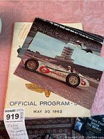 Photo showing the front cover of the 1962 Indianapolis 500 official program with two large color postcards on top, featuring the race car number 5 on the track.