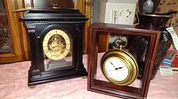 Photo showing two mantle clocks on a pink tablecloth, black wooden clock with mechanical movement and gold dial on left, suspended gold-tone pocket watch clock in wooden frame on right.