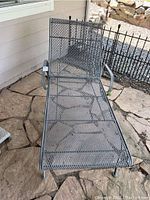 Full view of the metal chaise lounge on a stone patio showing its mesh surface and curved armrests.