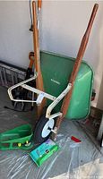 Metal and wood wheelbarrow with green basin and single front wheel, green hand held seed spreader, all placed on a plastic sheet in a garage.