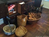 Photo showing woven baskets in varying shapes and sizes, two scarecrow decorations placed inside a large oval basket, part of wooden furniture visible in background.